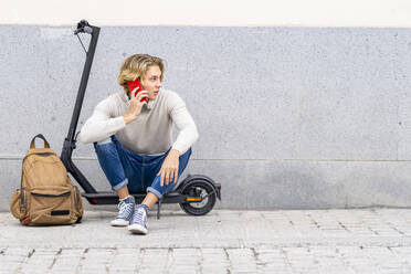 Man with backpack talking on mobile phone while sitting on electric push scooter against wall - GGGF00303