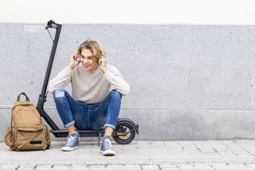 Smiling man with backpack wearing headphones while sitting on electric push scooter against wall - GGGF00304