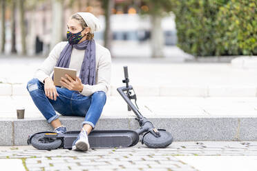 Mann mit Elektroroller und Gesichtsmaske, der ein digitales Tablet benutzt, während er auf einem Fußweg in der Stadt sitzt - GGGF00307