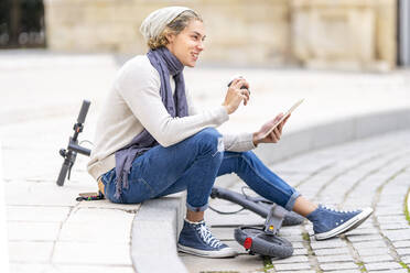 Smiling man with digital tablet drinking coffee while sitting by electric push scooter in city - GGGF00311