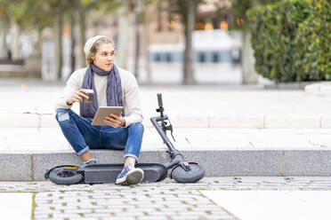 Mann mit Elektroroller, der auf dem Gehweg sitzend ein digitales Tablet benutzt - GGGF00312