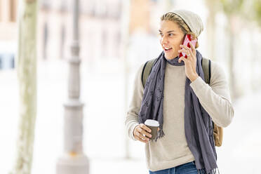 Lächelnder Mann mit Kaffeetasse, der beim Spazierengehen in der Stadt mit seinem Mobiltelefon spricht - GGGF00319