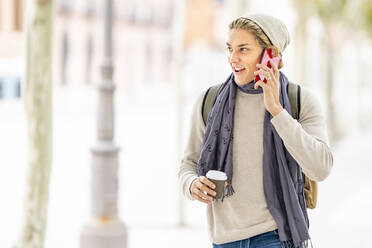 Smiling man with coffee cup talking on mobile phone while walking in city - GGGF00319