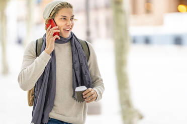 Junger Mann mit Kaffeetasse, der lächelt, während er mit einem Mobiltelefon in der Stadt spricht - GGGF00320
