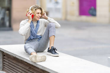 Young man listening music while sitting on bench in city - GGGF00336