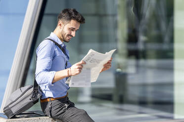 Businessman smiling while reading newspaper sitting outdoors - GGGF00347