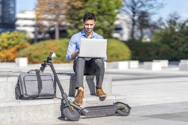 Businessman working on laptop while sitting by briefcase and electric push scooter in city - GGGF00349