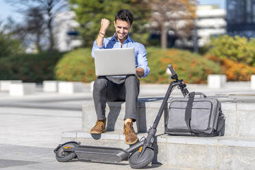 Smiling businessman showing winning gesture while sitting by briefcase and electric push scooter in city - GGGF00350