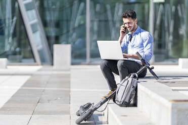 Lächelnder Geschäftsmann mit Laptop, der auf einem Mobiltelefon spricht, während er mit einer Aktentasche und einem Elektroroller auf einer Treppe sitzt - GGGF00351