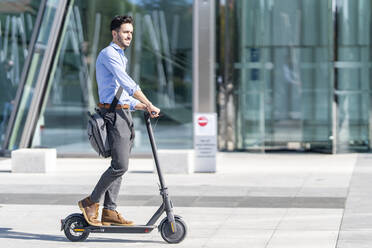 Young businessman with crossbody bag riding electric push scooter on footpath - GGGF00353