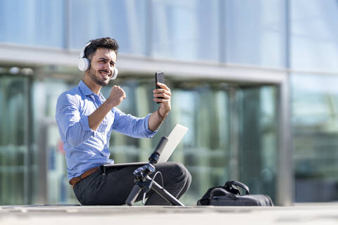 Lächelnder Geschäftsmann mit Kopfhörern, der eine gewinnende Geste zeigt, während er mit Laptop und Mobiltelefon im Freien sitzt, lizenzfreies Stockfoto