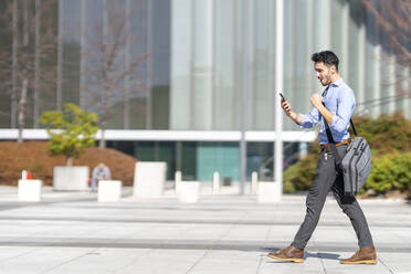 Young businessman with crossbody bag using mobile phone while walking on footpath in city - GGGF00356