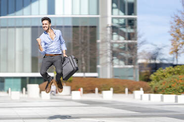 Excited businessman holding briefcase while jumping on footpath - GGGF00358