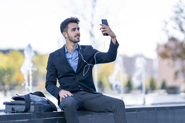 Geschäftsmann mit In-Ear-Kopfhörern, der ein Selfie mit seinem Mobiltelefon macht, während er auf einer Bank sitzt - GGGF00361