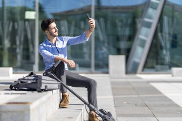 Businessman with push scooter and briefcase smiling while taking selfie sitting on steps - GGGF00363