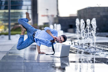 Businessman with laptop drinking coffee while doing handstand by fountain on footpath - GGGF00365