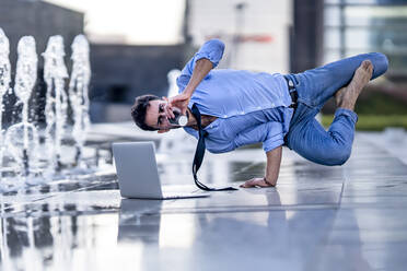 Young businessman with laptop drinking coffee while doing handstand by fountain on footpath - GGGF00366