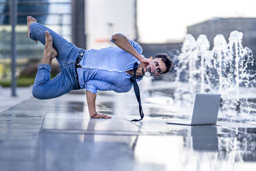 Businessman with laptop drinking coffee while doing handstand by fountain - GGGF00367