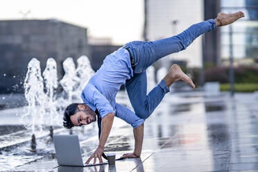 Businessman using laptop while doing handstand on footpath by fountain - GGGF00368