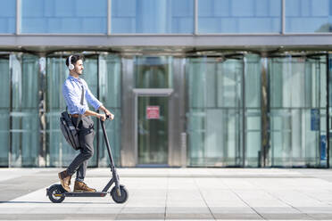 Junger Geschäftsmann mit Kopfhörern, der auf dem Gehweg einen Motorroller fährt - GGGF00371