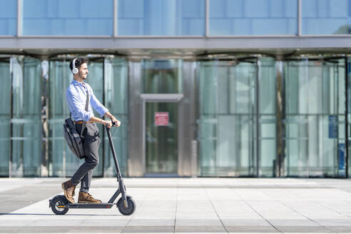 Junger Geschäftsmann mit Kopfhörern, der auf dem Gehweg einen Motorroller fährt - GGGF00371