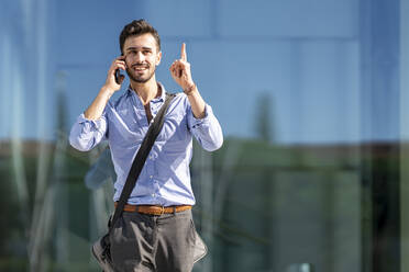 Businessman wearing crossbody bag pointing upward while talking on mobile phone outdoors - GGGF00372