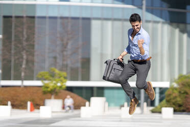 Young excited businessman holding briefcase while jumping outdoors - GGGF00373