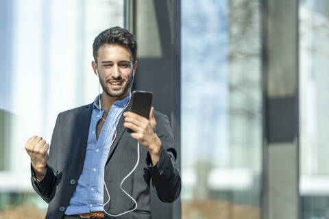 Smiling businessman wearing in-ear headphones cheering while using mobile phone standing outdoors stock photo