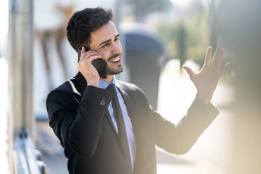 Lächelnder Geschäftsmann gestikuliert, während er im Freien mit einem Mobiltelefon spricht - GGGF00385