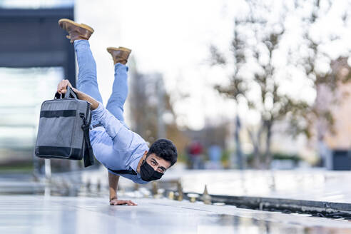 Man wearing face mask holding briefcase while doing handstand on footpath during Covid-19 - GGGF00386