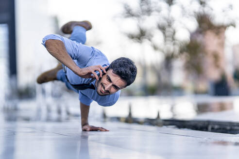 Smiling businessman talking on mobile phone while doing handstand on footpath - GGGF00387