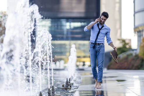 Smiling businessman talking on mobile phone holding shoe while walking barefoot by fountain - GGGF00390