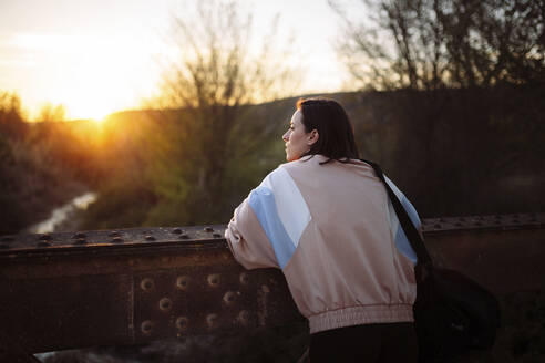 Frau betrachtet die Aussicht, während sie nach dem Training bei Sonnenuntergang auf einer Brücke steht - MRRF00725