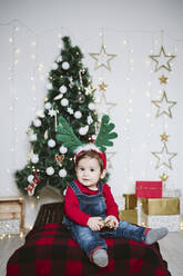 Niedlicher Junge mit gehörntem Stirnband, der an einem Weihnachtsbaum zu Hause sitzt - EBBF01695