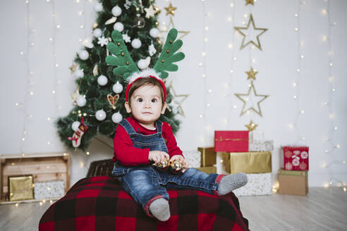 Kleiner Junge mit gehörntem Stirnband, der zu Hause an einem Weihnachtsbaum sitzt - EBBF01696