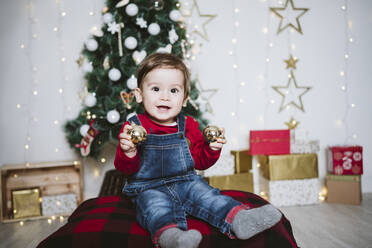 Glücklicher kleiner Junge, der mit einer Christbaumkugel spielt, während er zu Hause in der Weihnachtszeit sitzt - EBBF01697