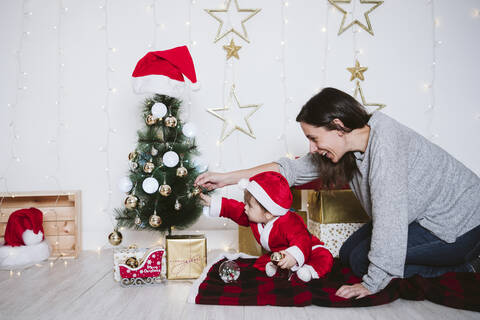 Mutter und Sohn spielen auf einer Decke sitzend mit einer Weihnachtskugel am Weihnachtsbaum, lizenzfreies Stockfoto