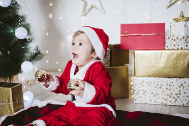 Verspielter kleiner Junge im Weihnachtsmannkostüm, der mit einer Weihnachtskugel spielt, während er zu Hause auf einer Decke sitzt - EBBF01703