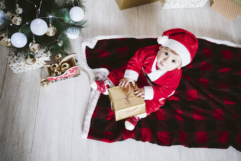 Kleiner Junge im Weihnachtsmannkostüm, der mit einem Geschenkkarton spielt, während er zu Hause in der Weihnachtszeit sitzt - EBBF01705
