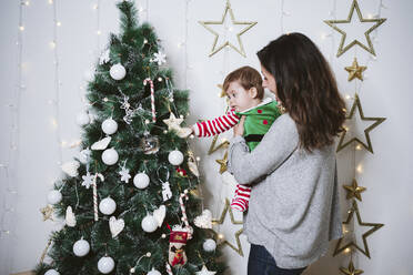 Mutter trägt ihren Sohn, während sie zu Hause am Weihnachtsbaum steht - EBBF01713