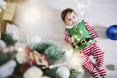 Kleiner Junge im Elfenkostüm, der mit einer Lichterkette spielt, während er in der Weihnachtszeit zu Hause auf dem Boden liegt - EBBF01718