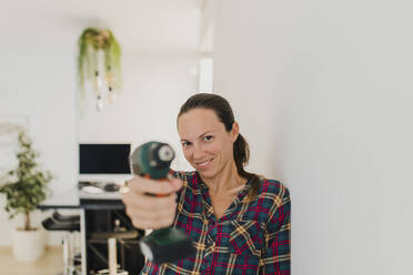 Lächelnde Frau zeigt elektrische Bohrmaschine, während sie sich zu Hause an die Wand lehnt - DMGF00404