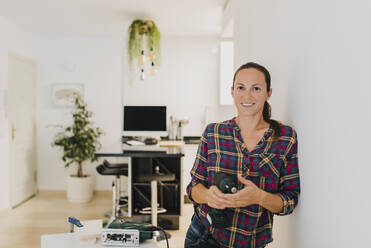 Mid adult woman holding electric drill while leaning on wall at home - DMGF00405