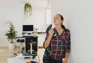 Woman holding electric drill while leaning on wall at home - DMGF00406