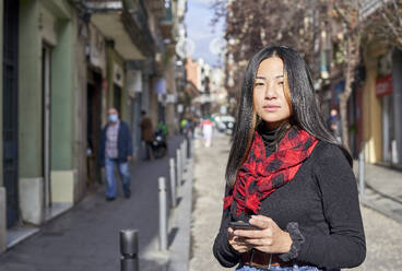 Schöne junge Frau, die ein Mobiltelefon benutzt, während sie auf der Straße in der Stadt steht - VEGF03233