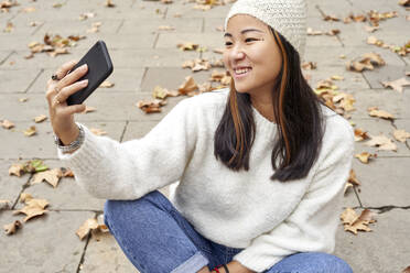 Lächelnde Frau, die einen Videoanruf auf einem Mobiltelefon betrachtet, während sie auf einem Fußweg sitzt - VEGF03244