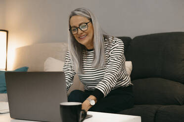 Smiling senior woman using laptop while sitting on sofa at home - ERRF04752