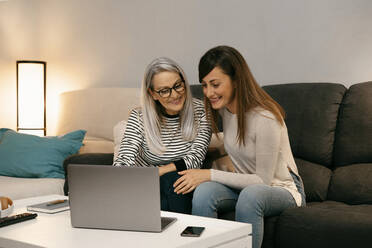 Smiling mother and daughter using laptop while sitting on sofa at home - ERRF04753