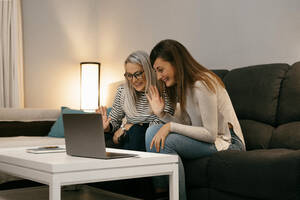 Lächelnde Frauen winken mit der Hand zum Videoanruf auf dem Laptop, während sie zu Hause auf dem Sofa sitzen - ERRF04754