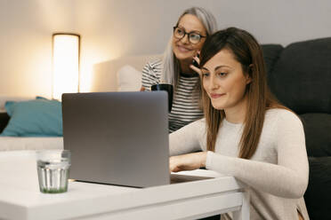 Daughter using laptop while mother talking on mobile phone sitting in background at home - ERRF04759